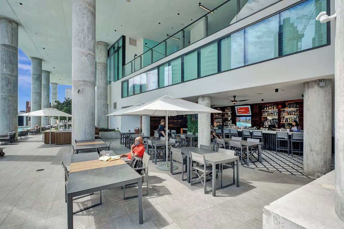 Un espace extérieur moderne, aménagé avec des tables et des parasols, un bar dont le menu est visible, et de grandes fenêtres offrant une vue sur le paysage urbain.