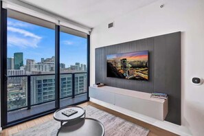 Ein modernes Wohnzimmer mit einem großen Fernseher an einer grauen Wand, einem kleinen runden Tisch und Blick auf eine Stadtlandschaft durch raumhohe Fenster.