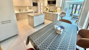 A modern kitchen with a central island, a dining area with a patterned tablecloth, and a view of the outdoors. - Modern Luxury Home - Hot Tub - 3 King Bedrooms - Free Parking - Close to Beaches (Poole)