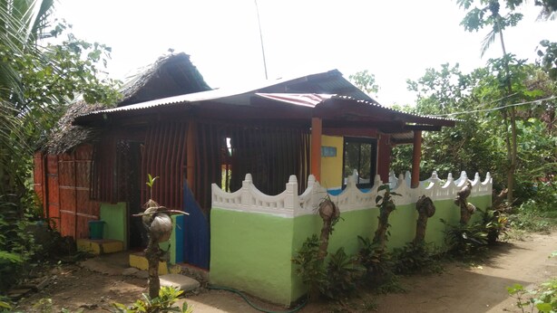 Una casa pequeña de un solo piso, con techo de lámina corrugada, entrada con portón y camino de tierra.