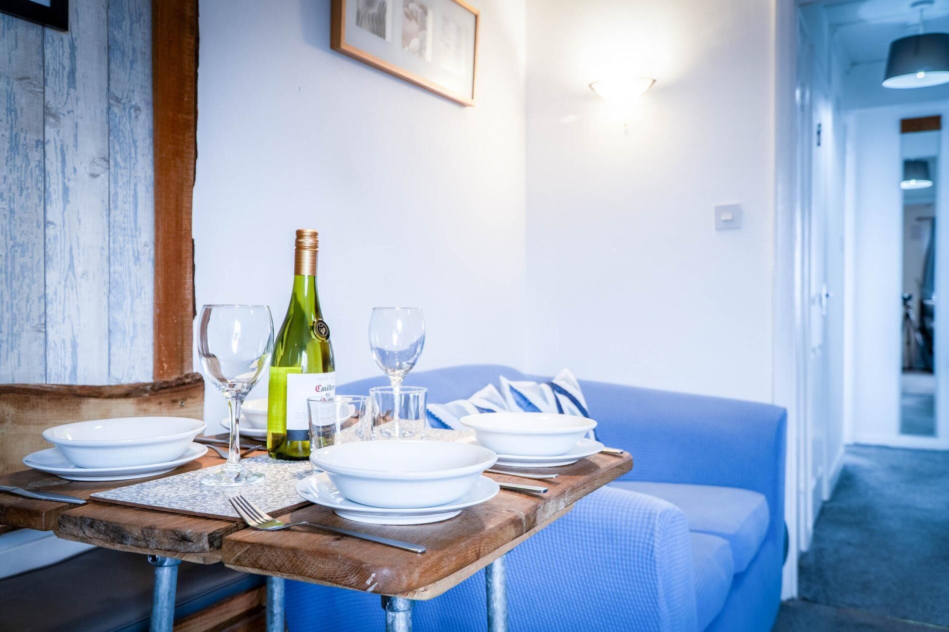 Una mesa de comedor dispuesta con platos, vasos y una botella de vino, con un sofá azul al fondo.
