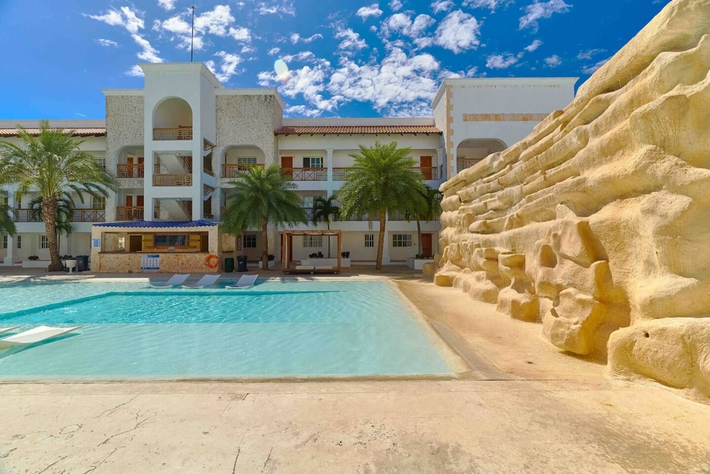 Ett hotell med pool, palmer och en klippig vägg.