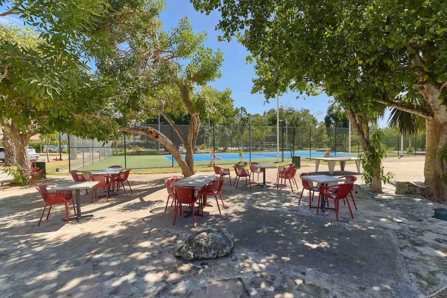 An outdoor seating area with tables and chairs, surrounded by trees and a fenced sports court in the background.