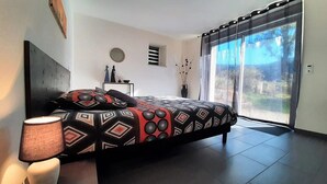Ein modernes Schlafzimmer mit einem großen Bett, einer Nachttischlampe, einem kleinen Tisch mit Dekoration und Blick durch eine Schiebetür.