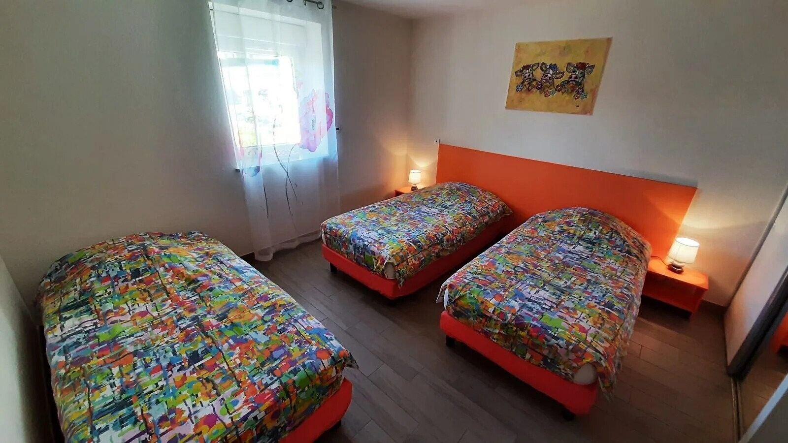 Ein Zimmer mit drei Betten, jedes mit bunter Bettwäsche, einem Fenster mit Vorhängen, einer Wand mit einem Gemälde und einem Nachttisch mit einer Lampe.