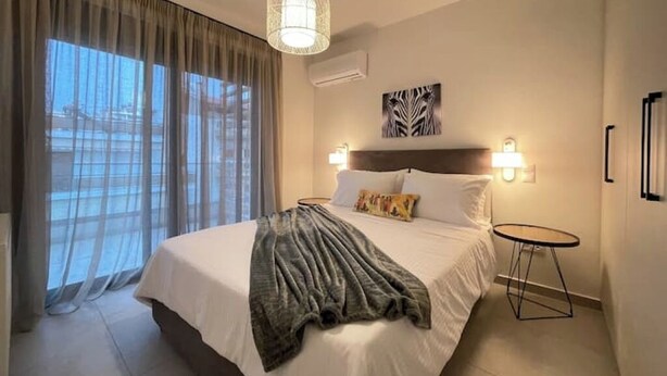 Una camera d'albergo con un letto grande, un comodino, una lampada a sospensione e una finestra con tende leggere.