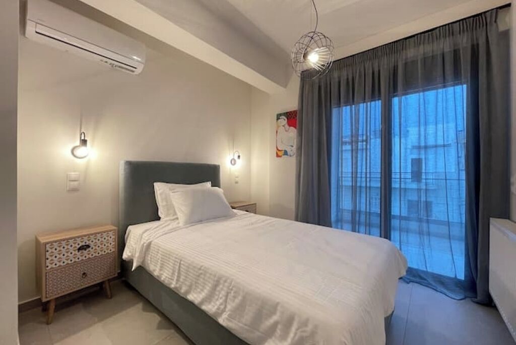 A bedroom with a bed, bedside tables, an air conditioner, a wall-mounted light, and a window with curtains.