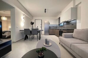 Ein modernes Wohnzimmer mit einer Couch, einem Couchtisch mit Pflanze, einem Essbereich mit Stühlen und Tisch sowie einer Küche mit Schränken und Geräten.