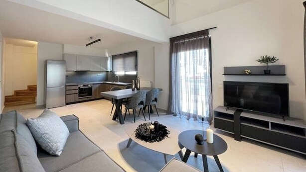 Uma sala de estar moderna com sofá, mesa de centro, lareira, televisão e cozinha ao fundo.