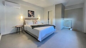 Quarto com cama, mesas de cabeceira, ar condicionado fixado na parede, um quadro e uma porta que dá para outro cômodo.