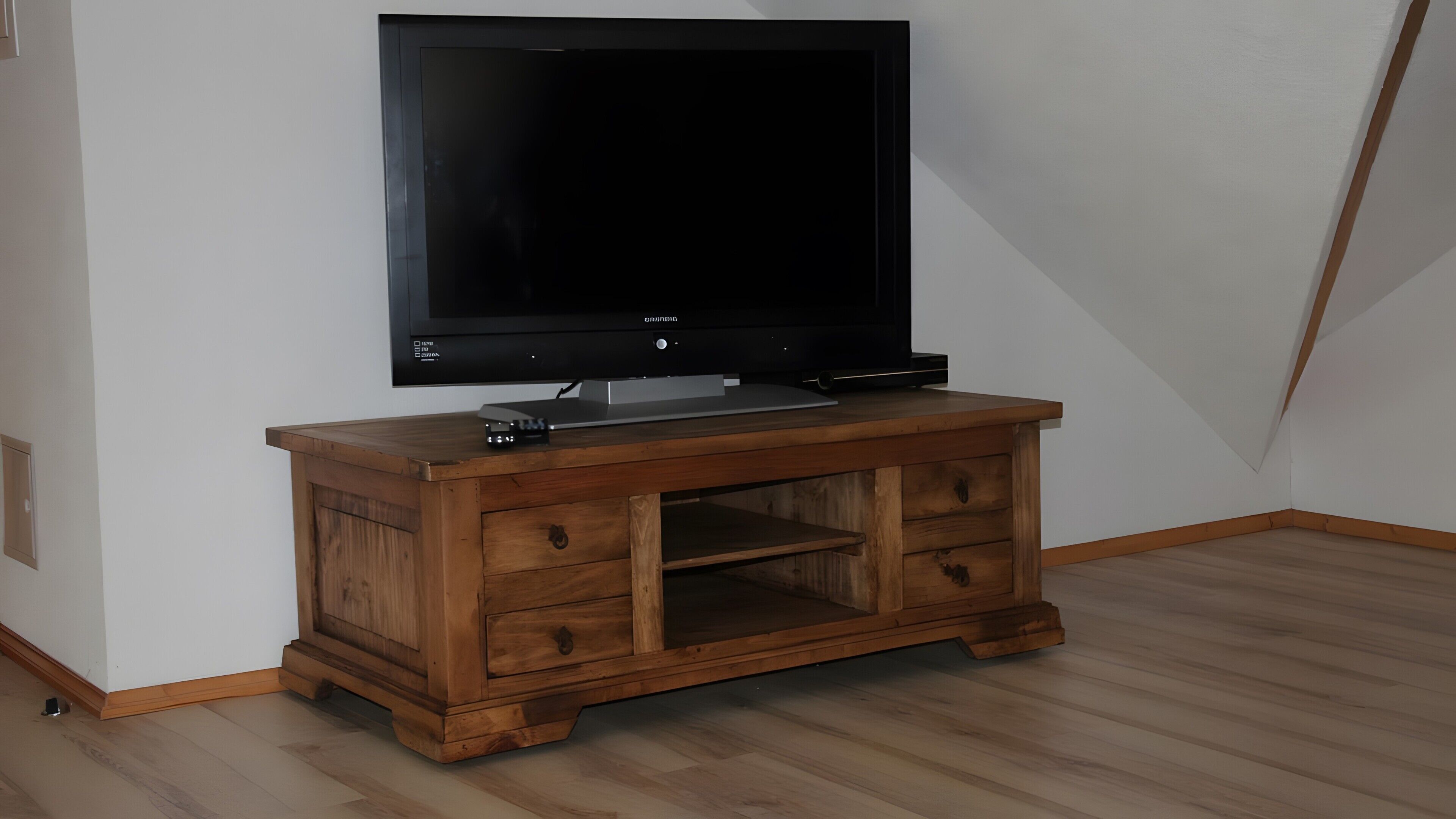 Huoneessa on puinen lattia ja valkoiset seinät, ja siellä on televisio, joka on sijoitettu puiseen jalustaan.