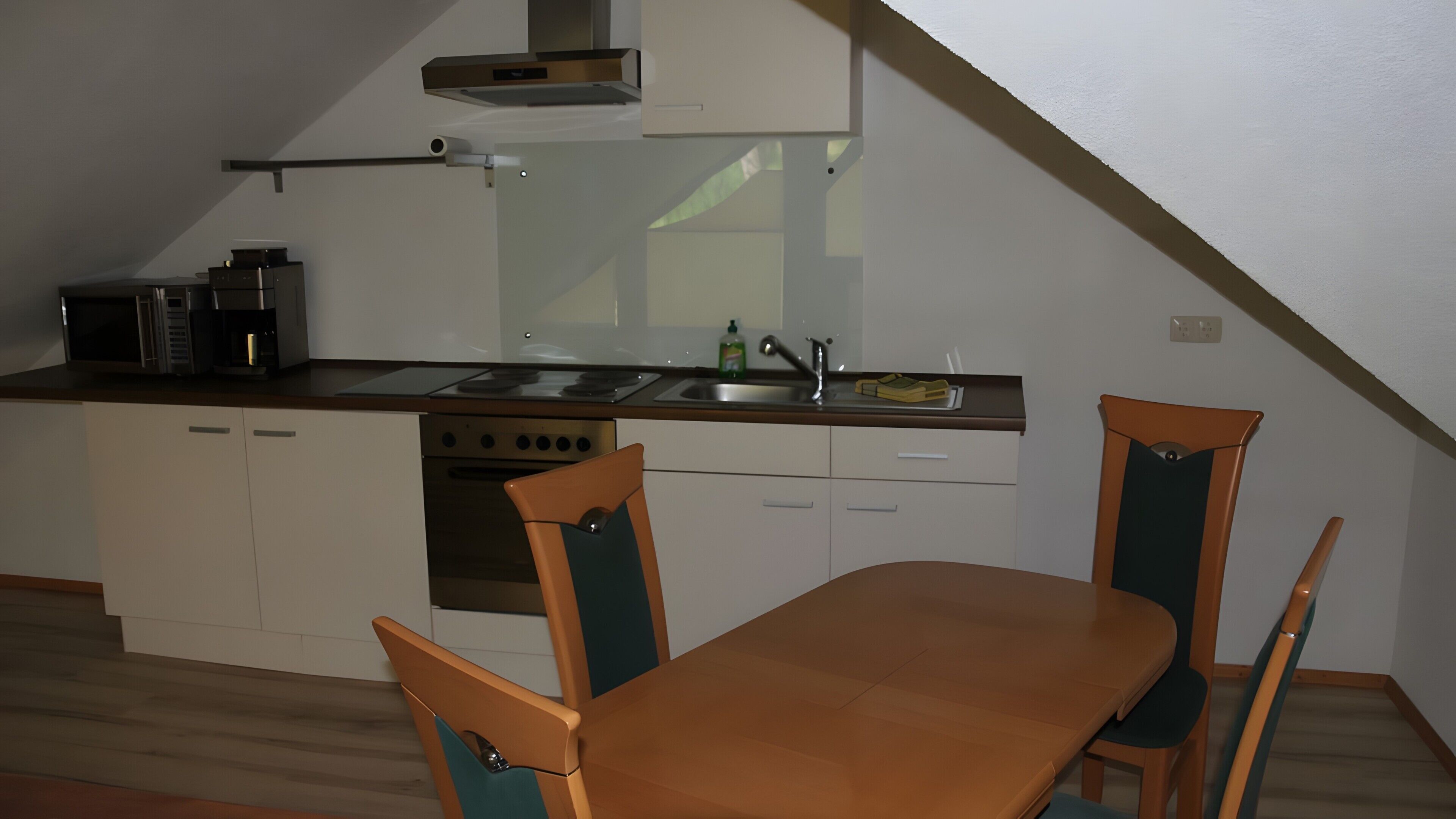 Una cocina compacta con estufa, lavamanos y armarios, una mesa de madera con sillas, y un techo inclinado.