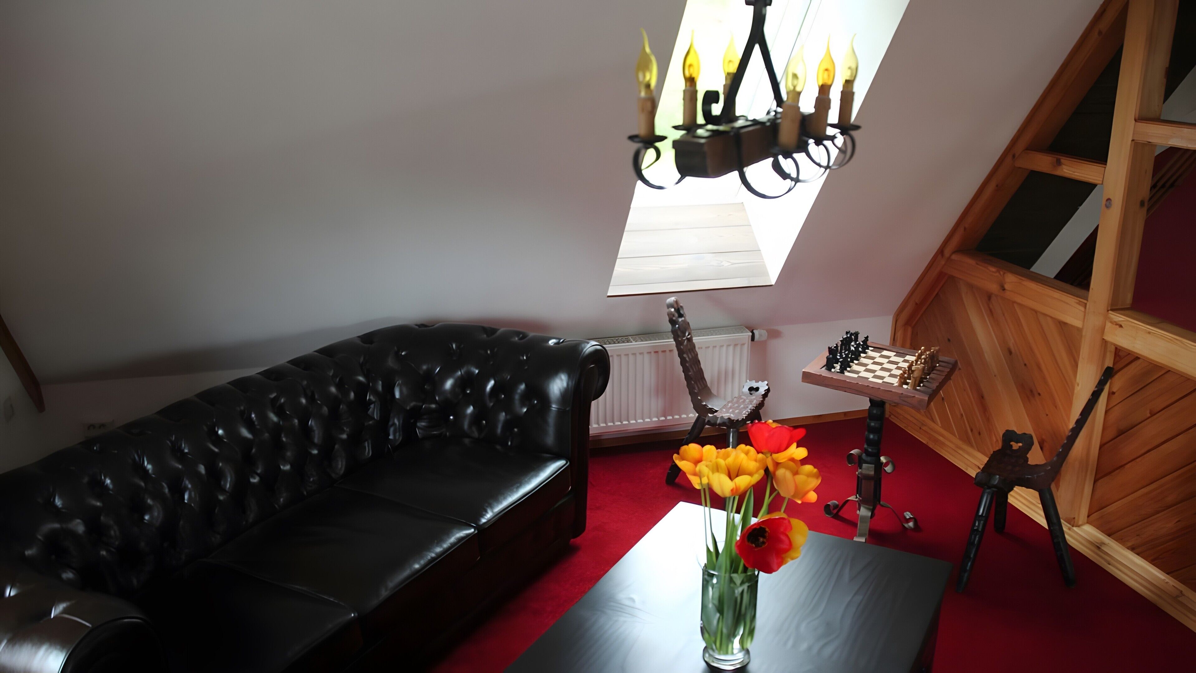 A room with a black leather sofa, a chessboard, a vase with flowers, and a chandelier.