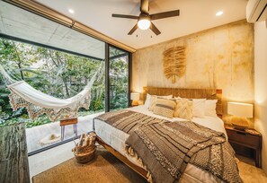 A bedroom with a large bed, a hammock, a view of greenery, a ceiling fan, and a wall-mounted lamp. - Stunning 2BR Jungle Oasis Tulum | Gym | Coworking (Tulum)