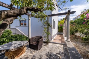 A covered outdoor seating area with a wicker chair, a stone bench, and a pathway leading to a white building with windows and shutters. - Luxury Garden Villa 50m from the Beach (Sutivan)