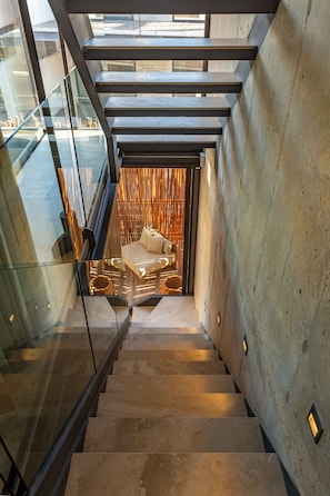 Un escalier moderne avec des rampes en verre, un coin salon avec des sièges rembourrés et un mur en béton.
