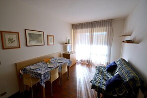 A dining area with a table set for four, a sofa, and a window with curtains. - Sunny Apartment Steps From the sea in Bibione (San Michele al Tagliamento)