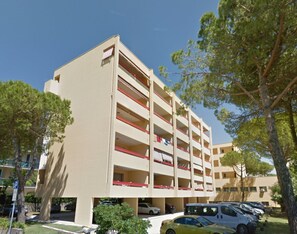 A multi-story building with a parking garage, trees, and parked cars. - Sunny Apartment Steps From the sea in Bibione (San Michele al Tagliamento)