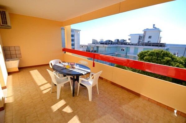 Un balcón con mesa y sillas, barandal rojo y vistas a edificios y vegetación.