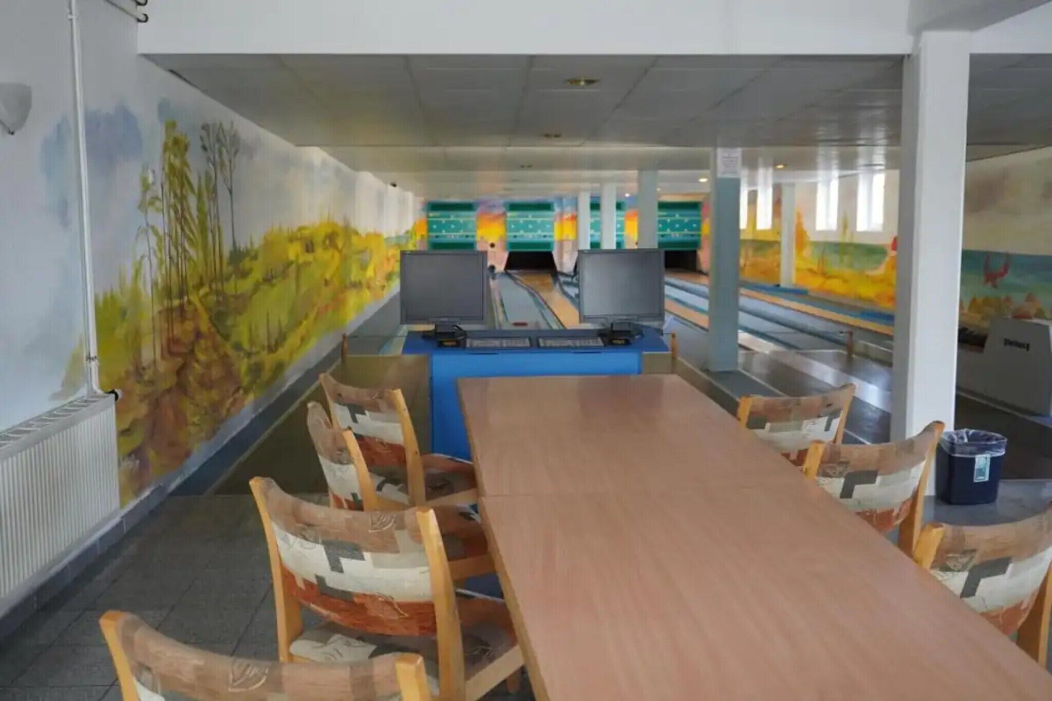 A bowling alley with a long wooden table and chairs, a bowling lane, and a mural on the wall.