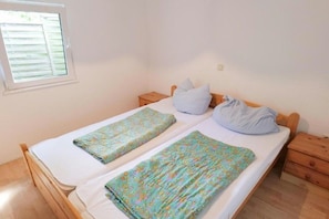 Ein Einzelbett mit weißer Matratze und zwei Kissen, ein Nachttisch aus Holz, ein Fenster mit Jalousien und ein Lichtschalter an der Wand.