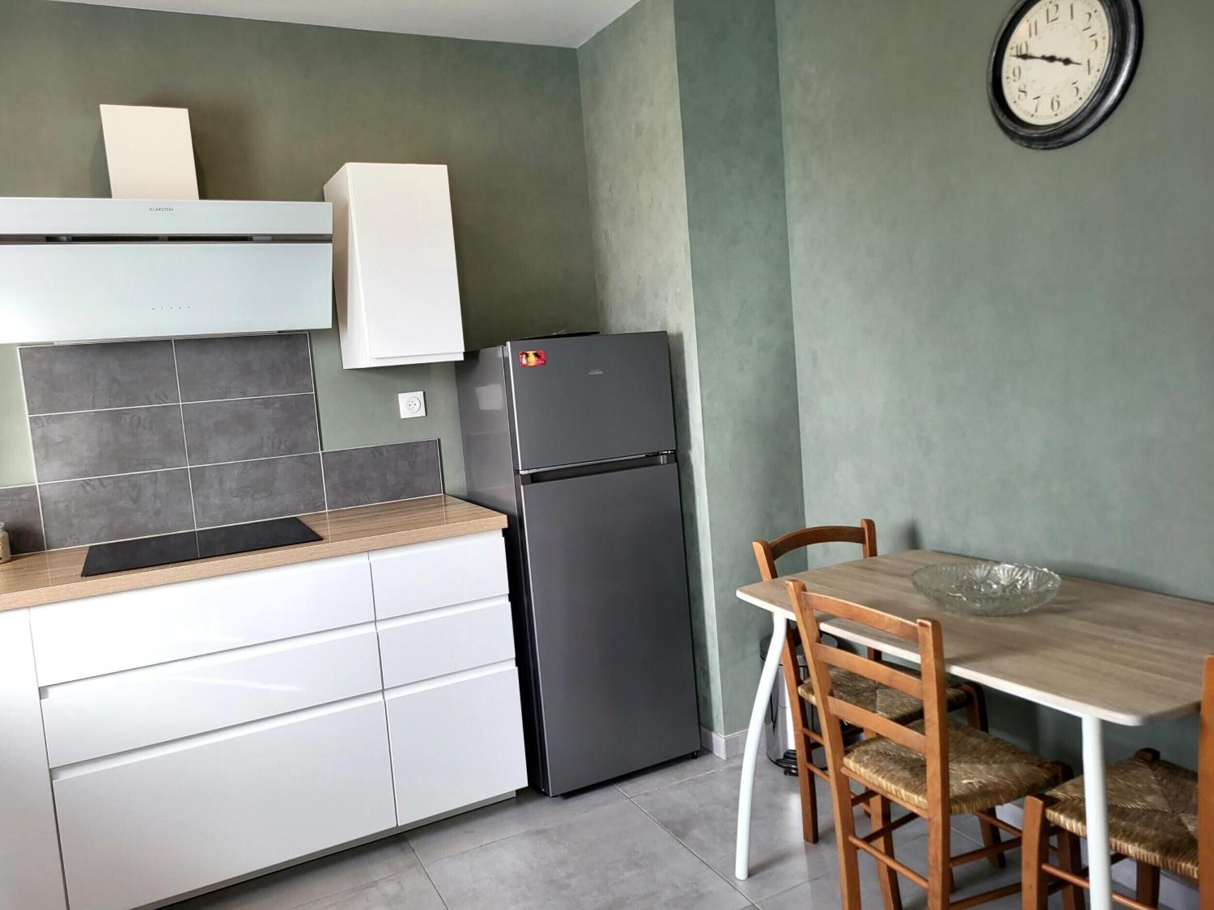 Une cuisine équipée d’un réfrigérateur, d’une table à manger avec des chaises et d’une horloge murale.