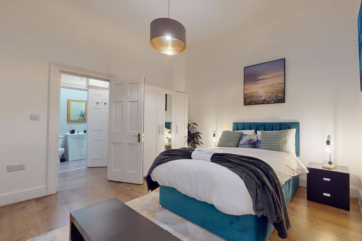 Un dormitorio con cama, mesita de noche, lámpara y un cuadro en la pared.
