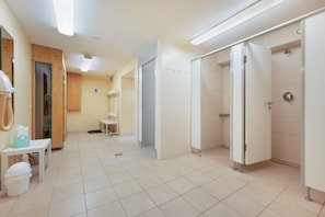 Un baño limpio y bien iluminado con azulejos blancos, zona de ducha y una pequeña mesa con una cesta y una botella.