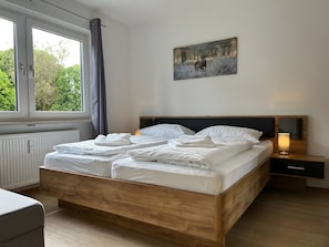 Ein ordentlich bezogenes Bett mit weißen Leinen, ein Holz-Kopfteil, ein Nachttisch mit Lampe und ein Bild an der Wand.