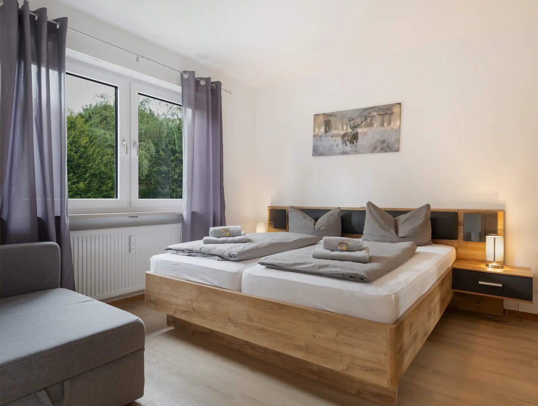 A bedroom with a wooden bed, a grey sofa, a window with curtains, and a painting on the wall.