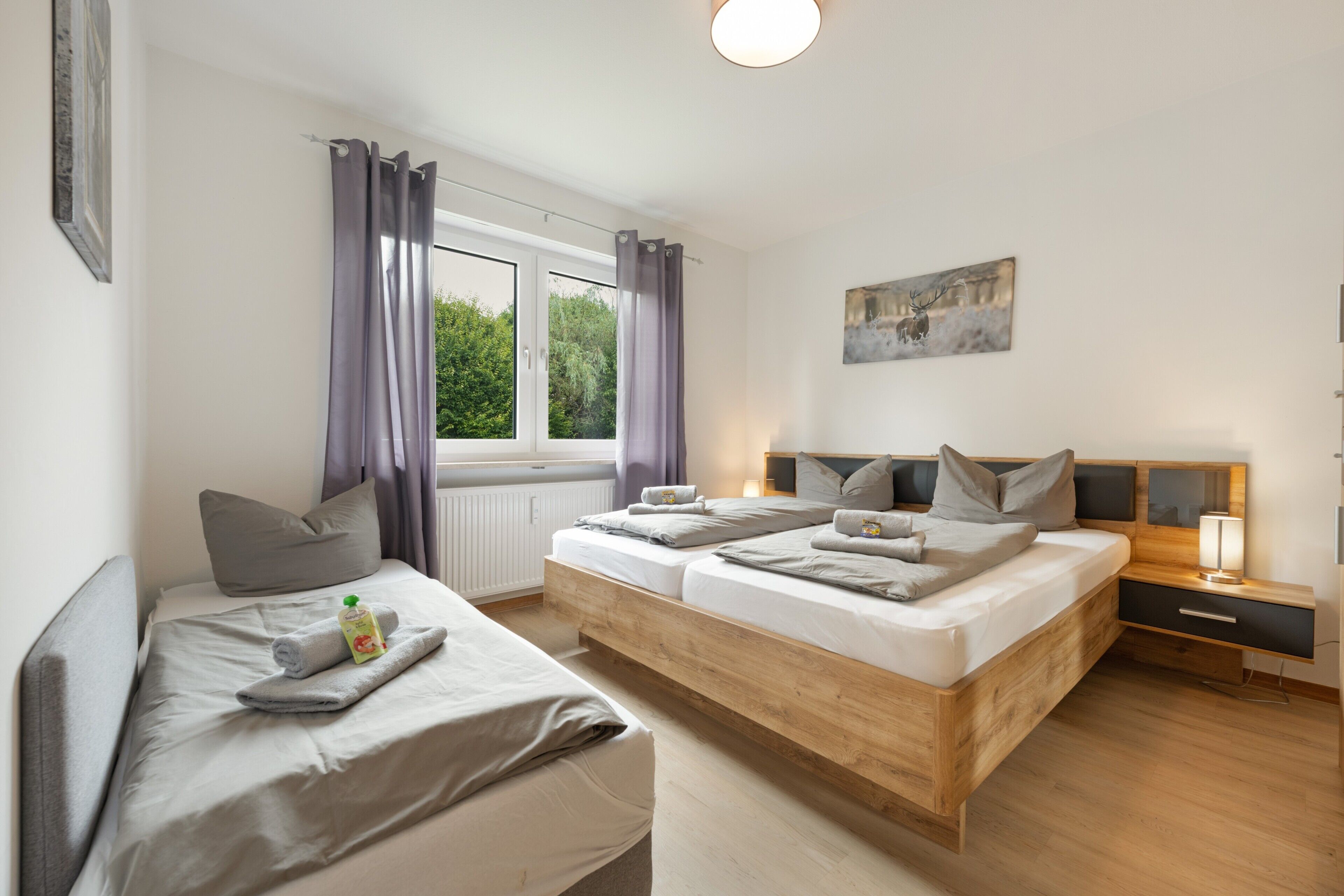 Ein modernes Schlafzimmer mit zwei Betten, einem Fenster mit Vorhängen, einem Nachttisch mit Lampe und einem Bild an der Wand.