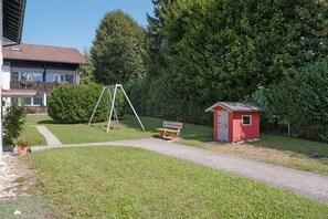 Un espacio verde con un columpio, un cobertizo rojo pequeño y un sendero que lleva a un edificio con balcón.