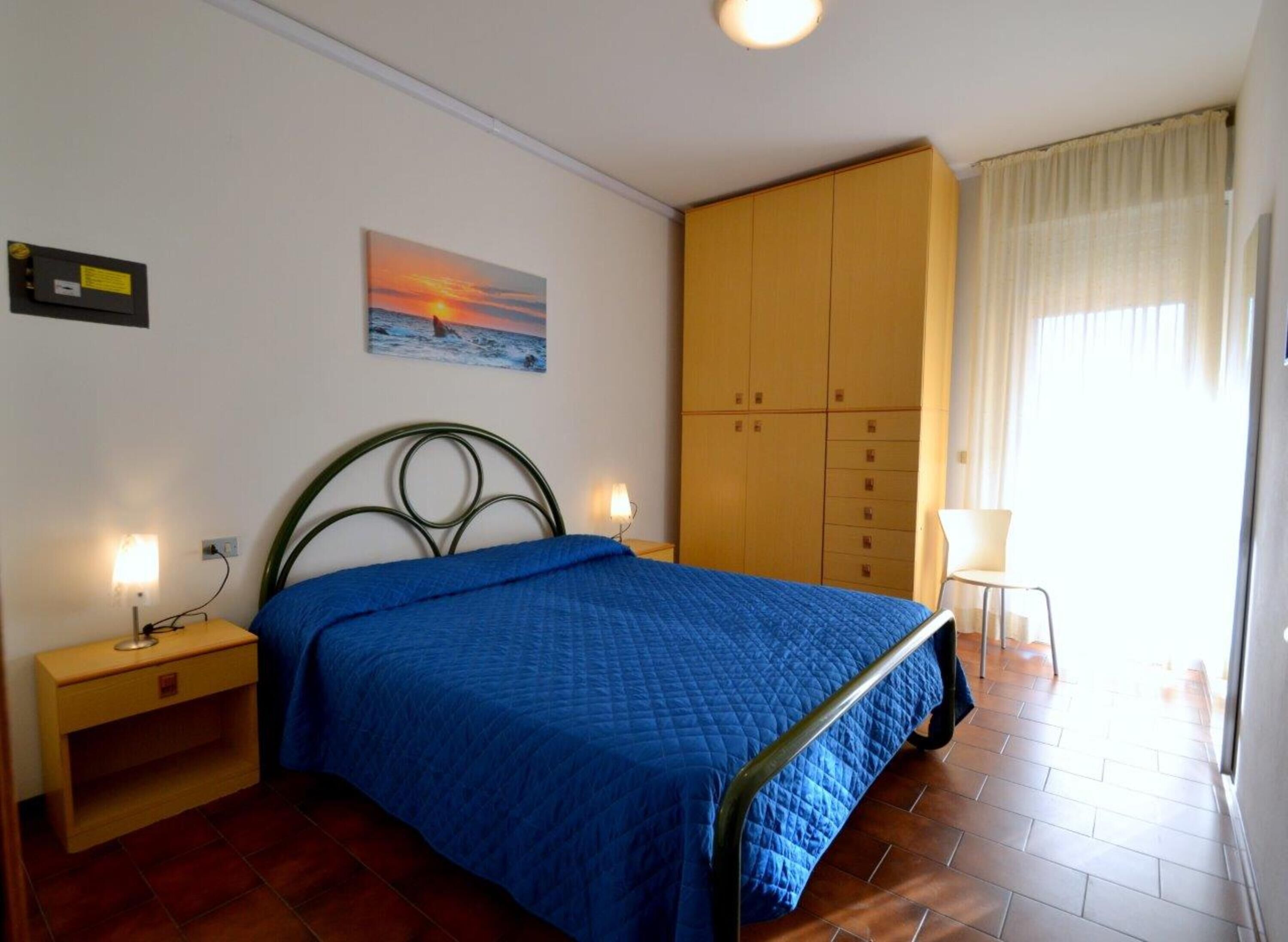 Un dormitorio con cama, mesita de noche, armario, un cuadro y ventana con cortinas.