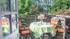 Een terras buiten met een gedekte tafel voor thee, omringd door stoelen met oranje kussens, en een houten hek op de achtergrond.