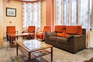 Un hall con un sofá marrón, sillas naranjas, una mesa de centro de cristal y cortinas estampadas.