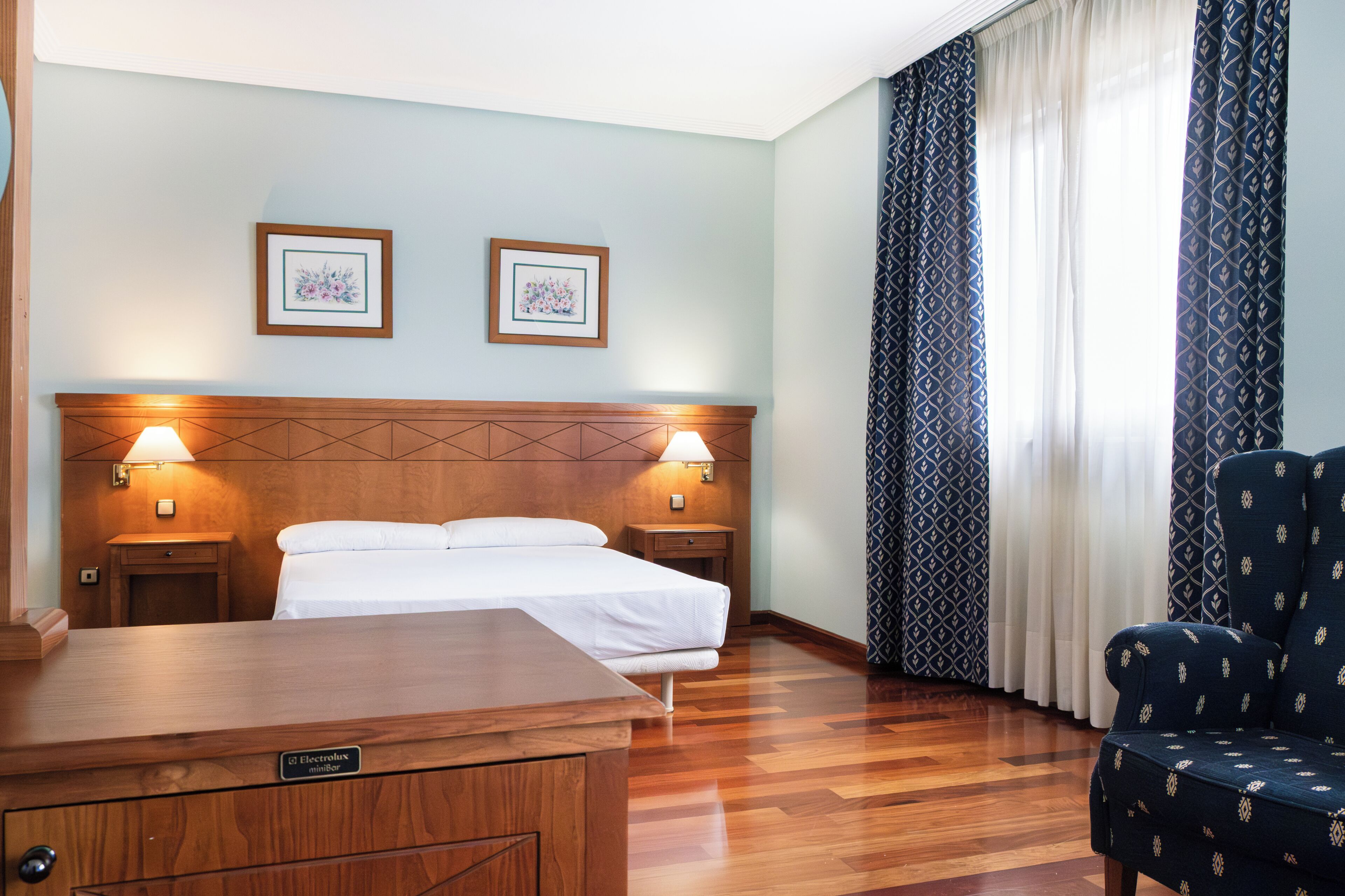 A hotel room with a wooden headboard, two bedside lamps, a bed with white linens, a desk with a nameplate, and patterned curtains.