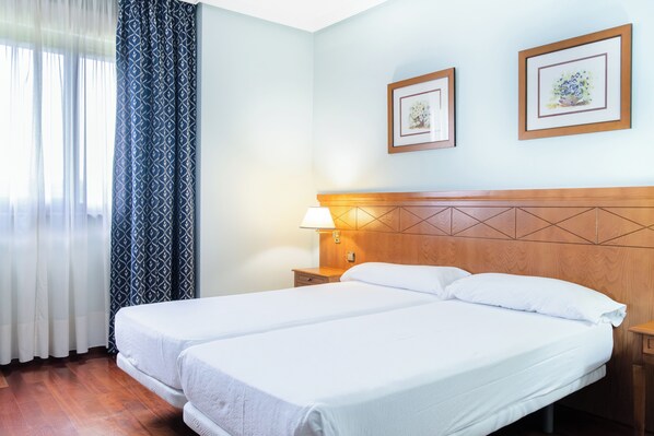 A hotel room with two beds, a wooden headboard, a nightstand with a lamp, and two framed pictures on the wall. - Checkin Basic León Norte (Villaquilambre)