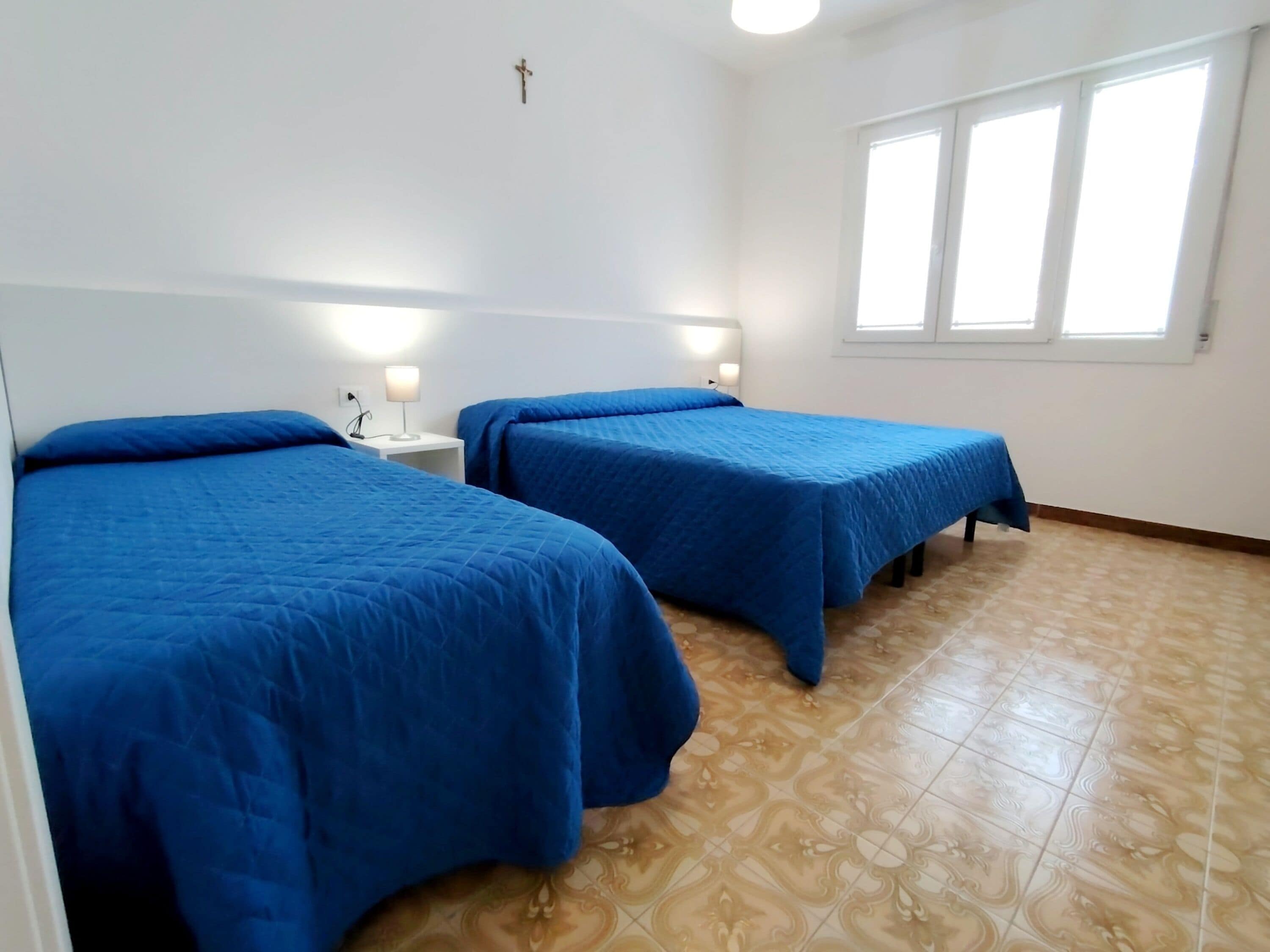 Una habitación con dos camas, una cruz en la pared y una ventana con persianas.