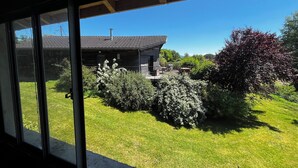 Blick durch ein Fenster auf ein Haus mit gepflegtem Rasen, eine Terrasse mit Gartenmöbeln und einem strahlend blauen Himmel.