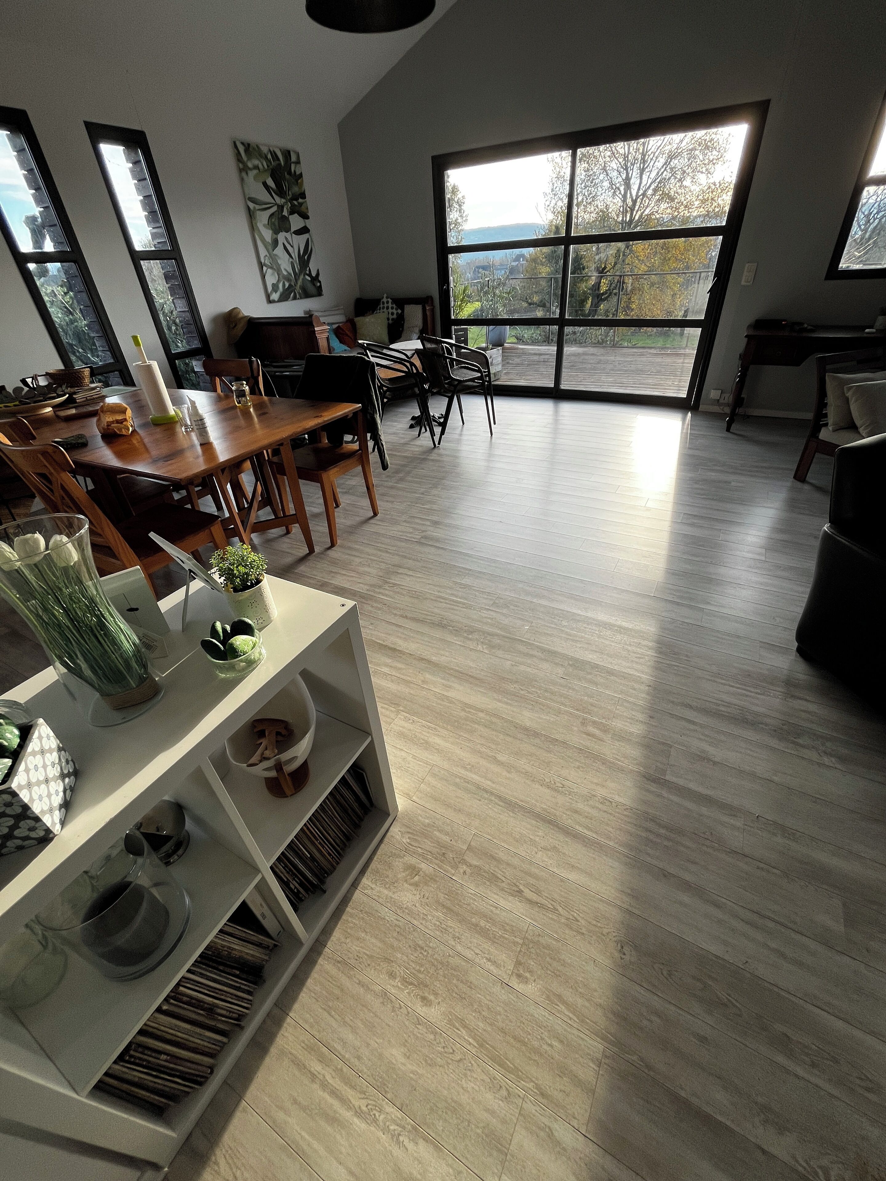 A spacious living area with a wooden floor, a dining table, chairs, a sofa, and a large window offering a view of the outdoors.