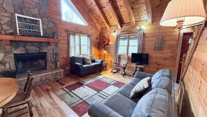 Un intérieur de chalet chaleureux avec une cheminée en pierre, des murs en bois, un grand canapé, un téléviseur et un coin repas.