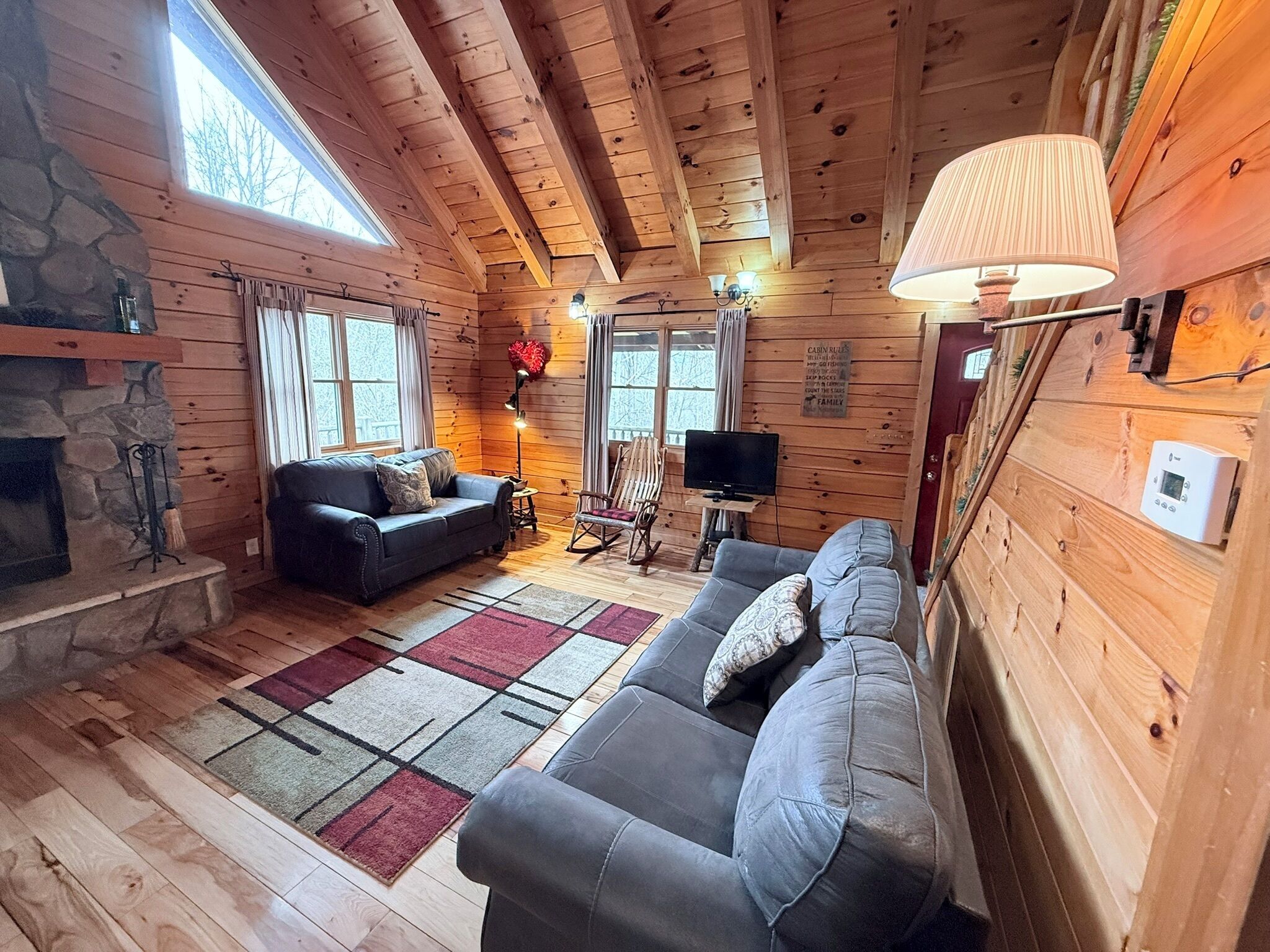 A cabin interior with a stone fireplace, two sofas, a television, and a large window.