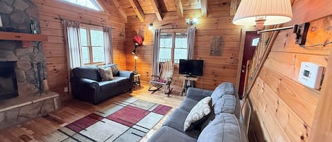 A cabin interior with a stone fireplace, two sofas, a television, and a large window.