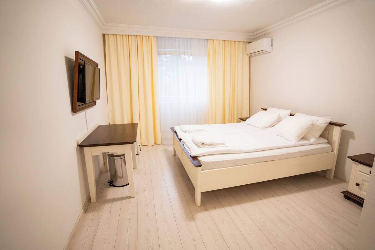 Une chambre à coucher avec un lit, un bureau, une poubelle et une fenêtre avec des rideaux.