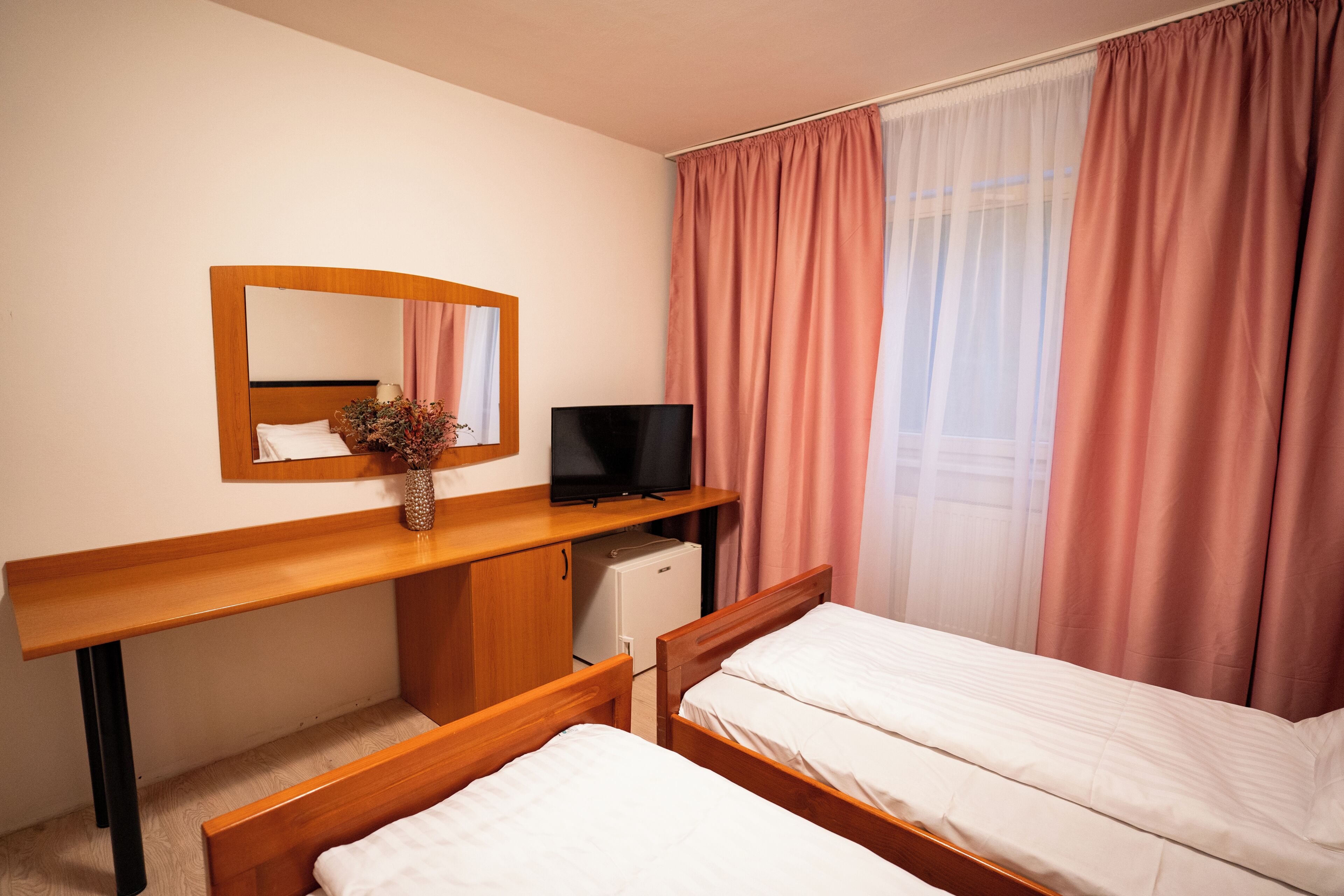 A hotel room with two beds, a TV, a desk, and a small refrigerator.