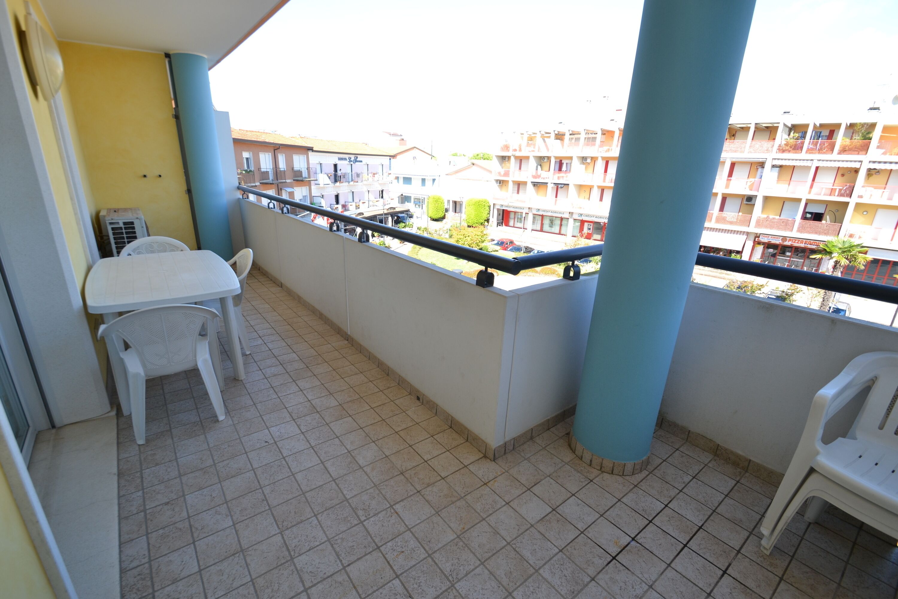 Een balkon met een witte tafel en stoelen, een betegelde vloer en een uitzicht op gebouwen op de achtergrond.