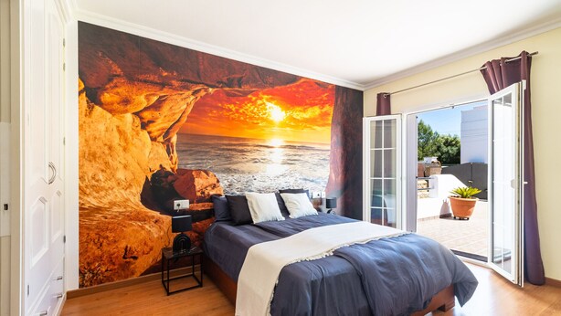 Un dormitorio con una cama, una mesita de noche, una lámpara y un mural con un atardecer sobre el océano.