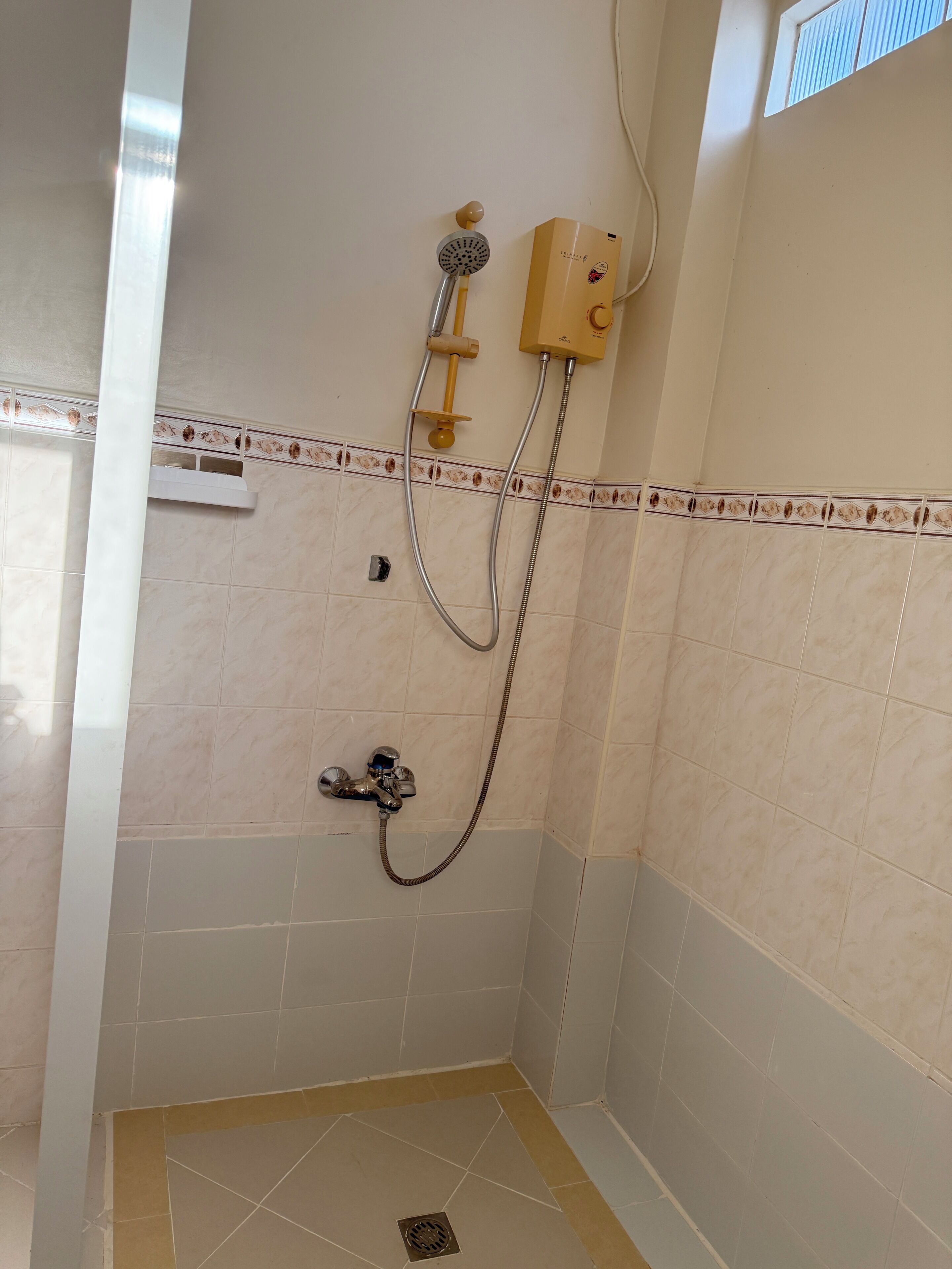 A small, tiled shower area with a handheld showerhead, a wall-mounted control unit, and a floor drain.