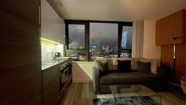 A modern living space with a kitchen, a sofa, a coffee table, and a large window with a city view. - Radio City Apartments near City Centre (Liverpool)