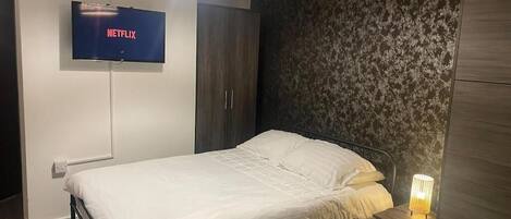 A bedroom with a bed, a TV mounted on the wall, a wardrobe, and a bedside lamp.